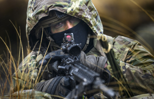 A soldier looks at the camera with a rifle in their hands, they are wearing a camouflage jacket with their hood up.