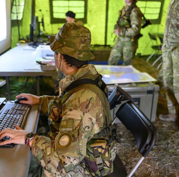 An aviator in a large green tent sits at a computer (left) while looking at a projected screen. Other aviators are watching as maps lay across an equipment case.
