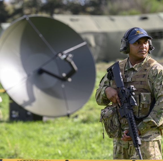 An aviator is full camouflage uniform, a blue RNZAF cap while holding a rifle stands in front of a satellite dish and tents as other aviators work in the background.