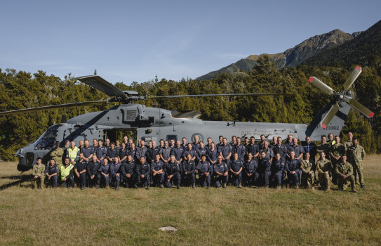 NZ Police staff and Royal New Zealand Air Force personnel stand in front of a large grey NH90 helicopter in front of a forrest and underneath blue skies.