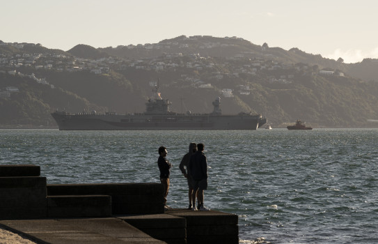 A large ship silhouette sits on the horizon as three people look on in the foreground. In the background, the hills of Wellington covered in homes stand tall.