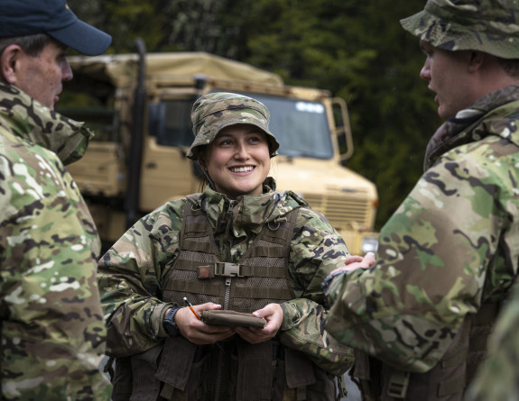 Three uniformed aviators stand with a unimog in the background. The two closest have their backs to the camera and the one with their face toward the camera is in the centre, smiling but looking at the aviator to the right. 