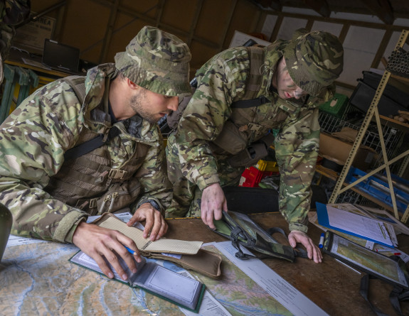 Two uniformed aviators are at a table - one is seated, one is standing up. They are looking at maps and have notebooks. They are inside somewhere. 