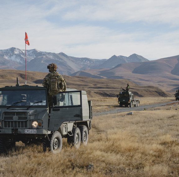 One small and three large Army vehicles on a road surrounded by dry grass, rolling hills and snow capped mountains in the background. A soldier stands above the vehicle in the front - it also has a red flag attached to the roof.