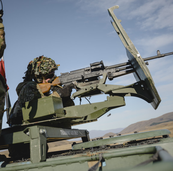 A soldier fires a weapon from the top of a MHOV in front of blue skies.