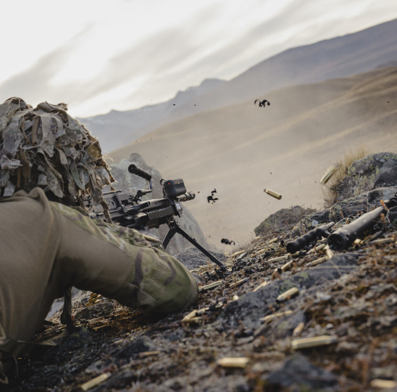 A soldier in the prone position on the floor, fires rounds down range as shells exit the weapon.