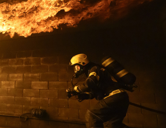 A person stands in fire fighting gear with flames above their head.