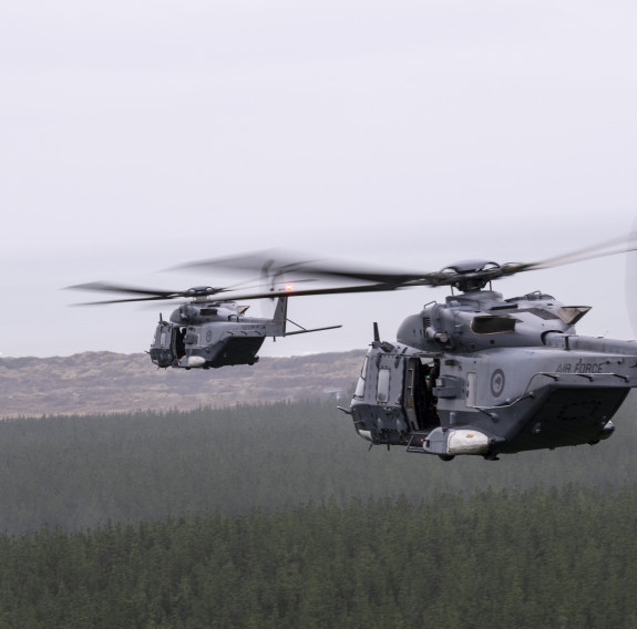 Two large dark grey helicopters fly in formation over a pine forest under grey clouds.
