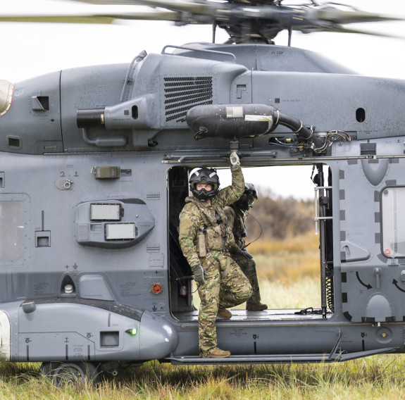 With the NH90 helicopter a loadmaster stands on the exterior step, under the side door of the helicopter. They have one hand up holding on the frame. The person takes up less than half of the open door space and the helicopter fills the frame.
