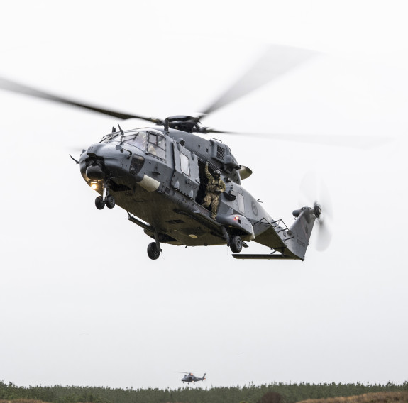A loadmaster stands at the open side-door of an RNZAF NH90 helicopter as it comes into land. In the distance is a RNZAF A109 helicopter flying above the trees under a sky full of cloud.