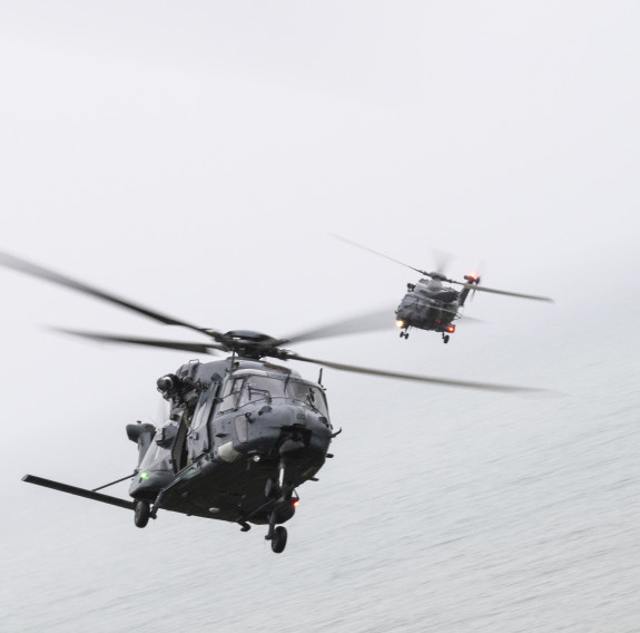 Two NH90 helicopters bank to the left, on in front of the other, over the ocean under great clouds.