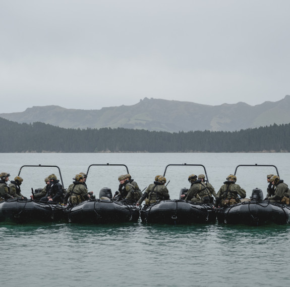 Four boats together with soldiers on board on still water in front of layered hills in the background.