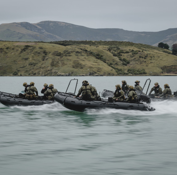 Three small boats of soldiers moves fast over the water, the background is blurred with motion.