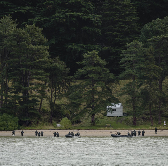 In the distance across the water, two boats can be seen unloading soldiers on a beach with trees and a large hill in the background.