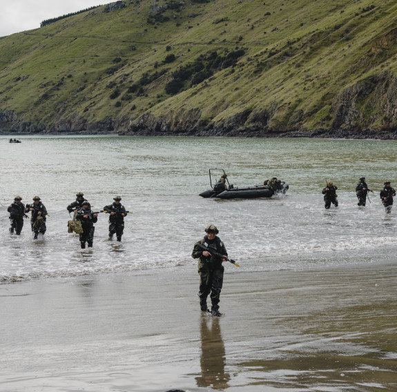 Two Zodiac boats sit just off the shore and soldiers make their way onto the beach. The soldiers are in wet weather gear and each wearing helmets and holding weapons.