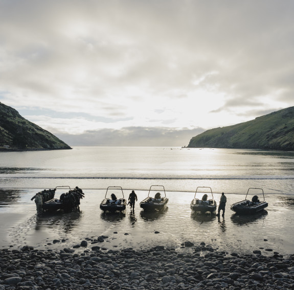 Four boats sit on the beach under harsh sunlight in a fjord as soldiers carry one of the boats up the beach.