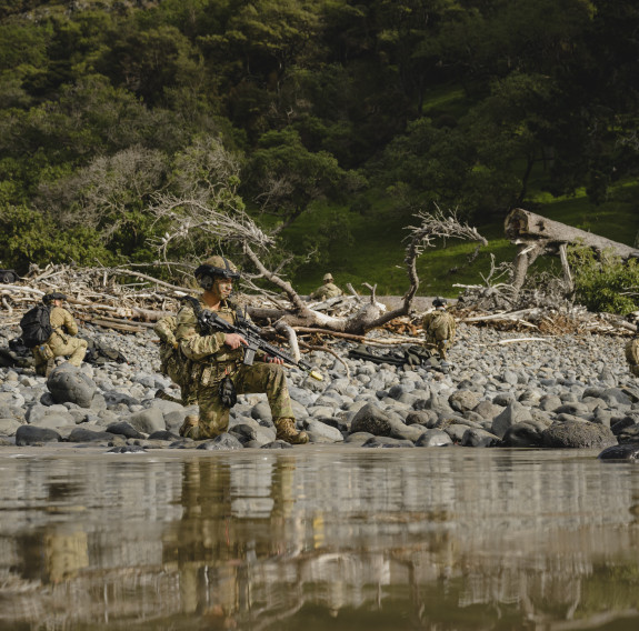 Soldiers are spread out and crouched down by the waters edge amongst rocks and driftwood. In the background are trees that cover a hill. the light is getting lower in the sky.