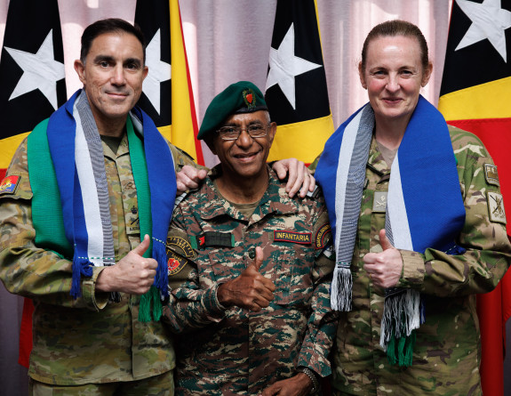 Three uniformed military personnel stand smiling and looking at the camera with their thumbs up. They are standing in front of the Timor-Leste flag. 