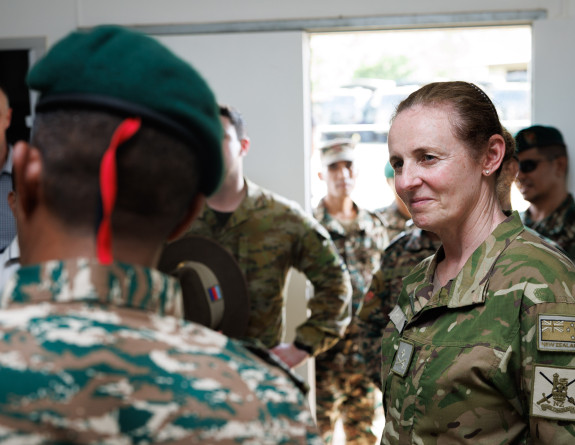 A uniformed NZ Army soldier looks at another uniformed military person with their back to the camera. In the background, there are other people in uniform looking toward this engagement .