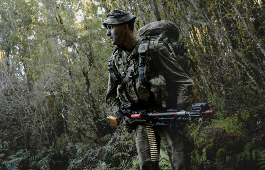 A soldier walks through dense bush fully equipped with a weapon and pack.