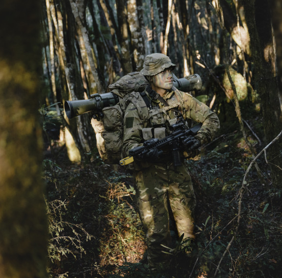 A soldier in full camouflage uniform and cam paint walks through dense forest with a full pack on.