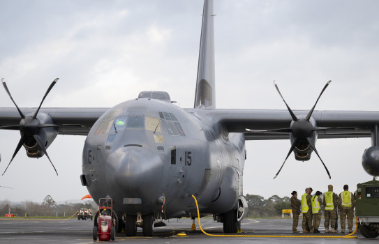 A large grey aircraft with propellors with four blades each sits on the tarmac as ground personnel huddle to the right. Towards the nose of of the aircraft is the number 15.