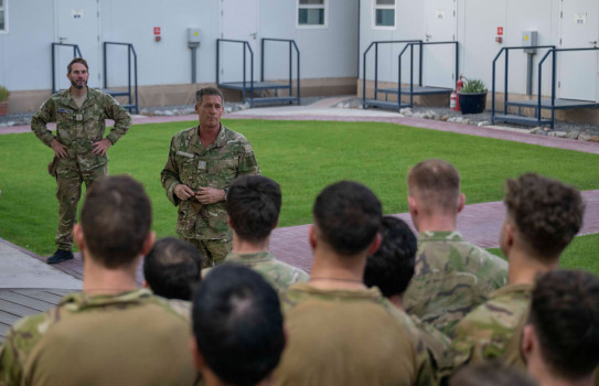 Two soldiers face the camera while the backs of at least 10 soldiers heads can be seen facing them. In the background is a square courtyard with lush green grass and steps into buildings.