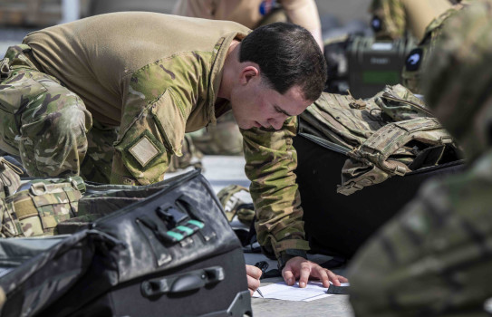 A soldier writes on a form on the ground surrounded by large packs and bags.