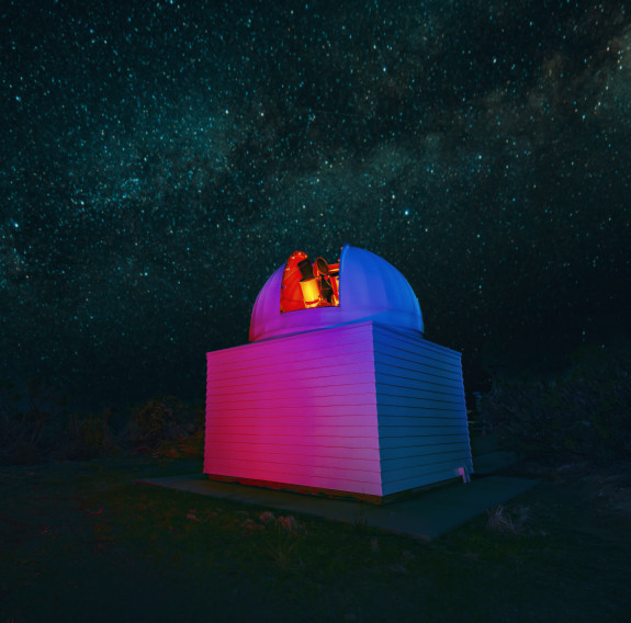The Defence Science Technology telescope sits under the stars. The outer dome opens across the centre to reveal a glowing red telescope. The outside of the building, usually white, is illuminated with red and blue lights.
