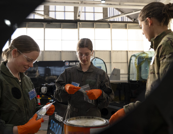 Inside a hanger, two students in dark green overalls, wearing protective glasses and orange gloves look at a small plastic bag they are each holding as an instructor looks on. 