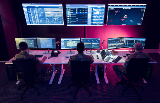 Inside No.62 Squadron at Base Auckland. Three large screens are mounted to the while and in front of them are three desks, each operated by an aviator in front of dual screens as red light glows across the room.