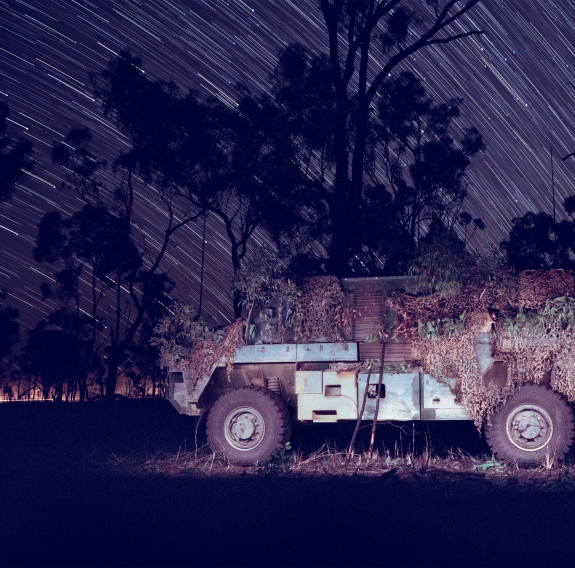 An Armoured Bushmaster Vehicle covered in camouflage is parked next to trees under the night sky. The long exposure photography creates star trails in the sky.