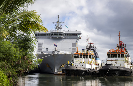 Framed by trees on the left, a large great ship has its bow facing the camera as it's tied up at a wharf accompanied by two tug boats ion the right,