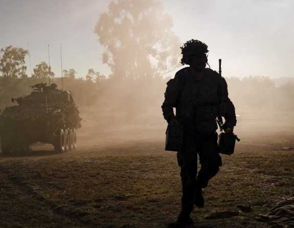 A silhouette of a soldier running towards the camera holding ammo containers with a Light Armoured Vehicle in the foreground and tree is the background. It is soft, evening light with dust in the air.
