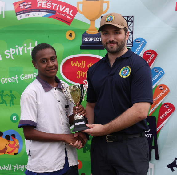 A young boy on the left and LT Cunningham in a polo and cap on the right hold a trophy and shake hands in front of a large event map.