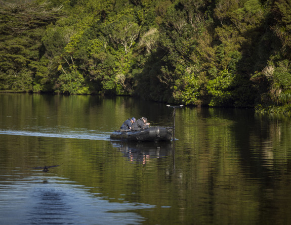 Navy hydrographers in a small black boat scan what's under the water with equipment mounted to the side of the dinghy.