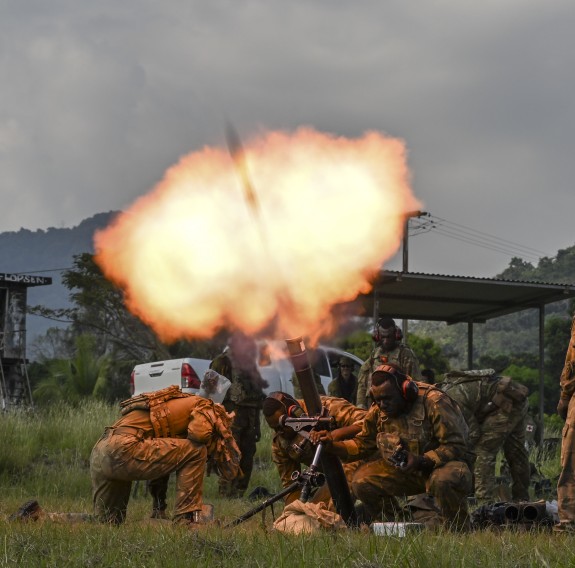 Soldiers fire a mortar that creates a fireball as the round leaves the barrel. Soldiers are illuminated by the flare as they crouch down towards the grass under cloudy skies.