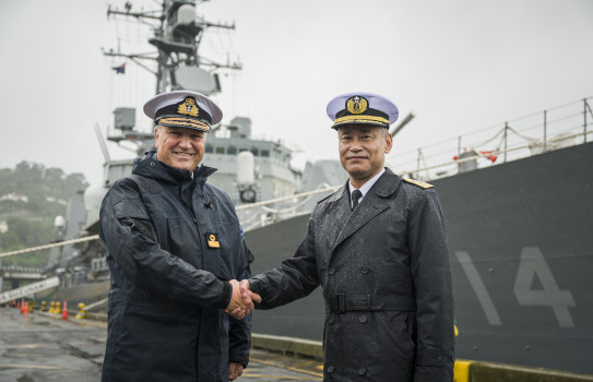 Two men shake hands in front of a large grey ship.