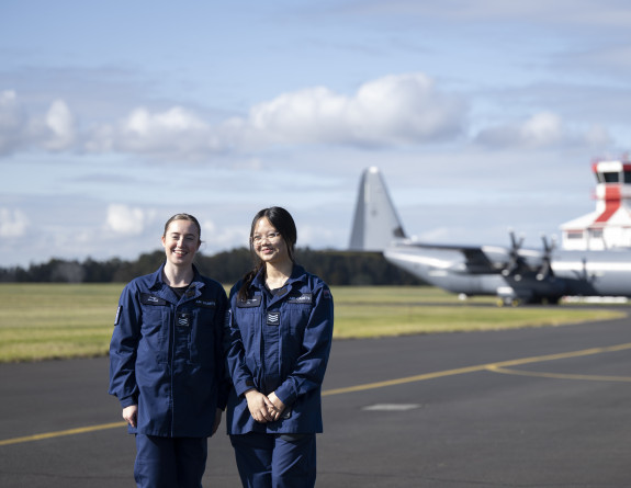 Two cadets on the tarmac in front of a large grey aircraft, the C-130J Hercules and the control tower painted in white and red.