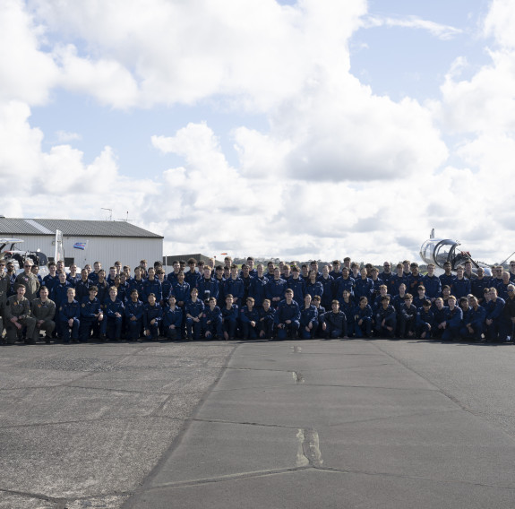 In front of two aircraft and a hanger, over 100 cadets pose for a photo with the RNZAF student pilots under scattered clouds.