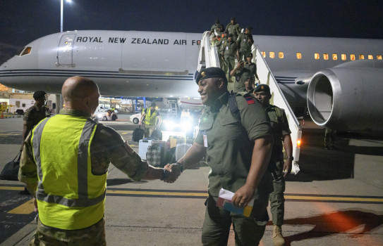 Two people shake hands after disembarking from a large grey aircraft that has the words "Royal New Zealand Air Force" written down the side. 