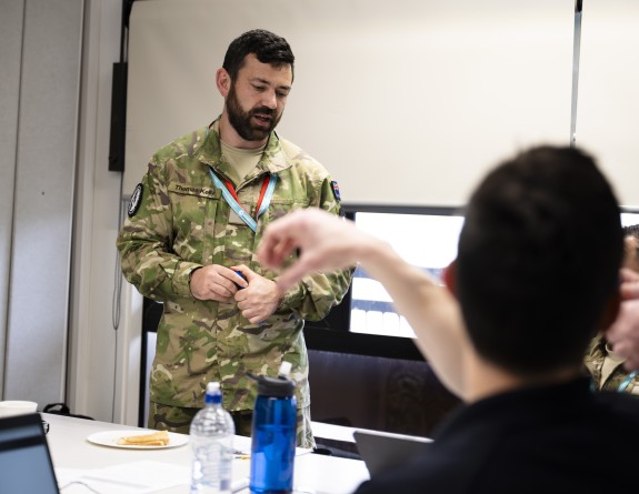 Personnel talk in a classroom, one person standing in military uniform and others out of focus in the foreground.