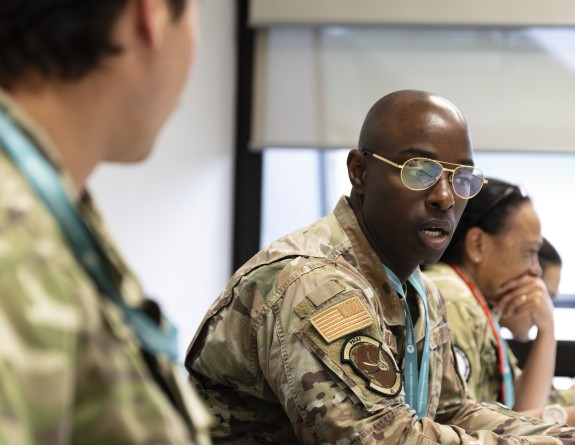Three military personnel sit at a table in a classroom setting while having a discussion.