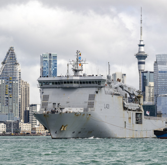 A large grey ship, HMNZS Canterbury and a blue and green tug, sail in front of the Auckland City skyline, with the iconic Skytower in the background under cloudy skies,