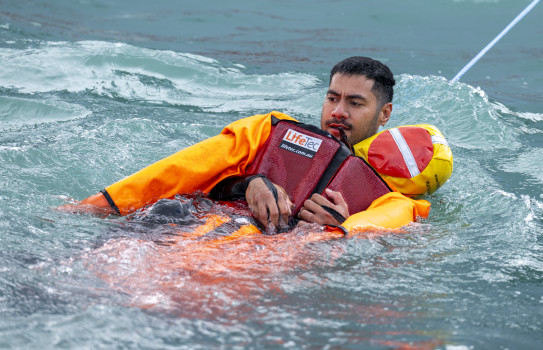 A sailor swims on his back in the water, holding a yellow and red mannequin under the arms. The sailor has a rope attached to safety from his back.