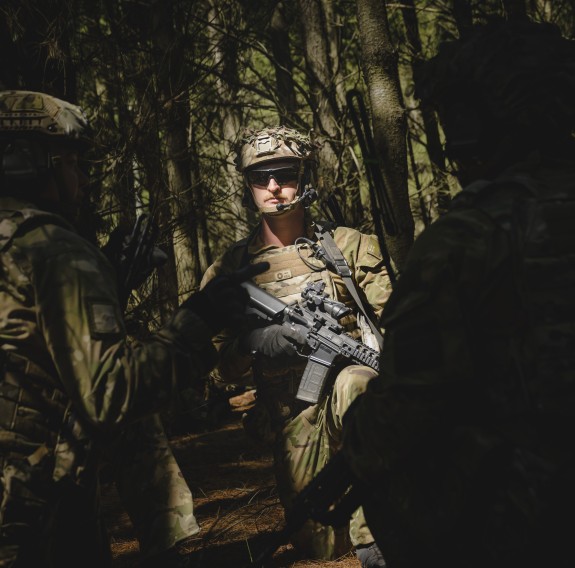 Soldier stand in a forest as the light through the trees lights a soldiers face.