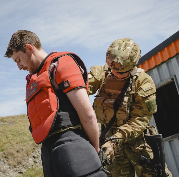 A soldier in Army uniform detains another soldier, wearing a red padded vest, who is acting as the enemy.