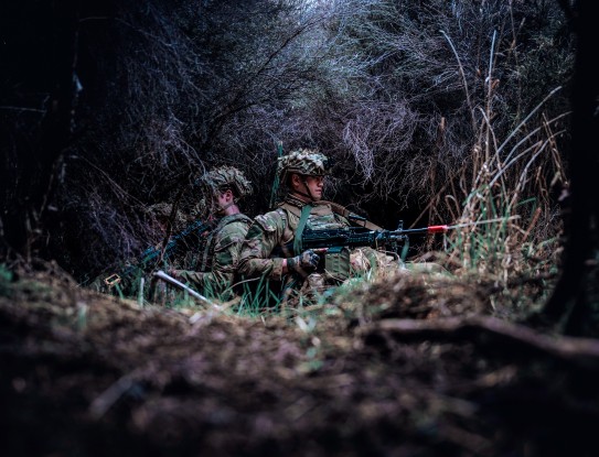 Two New Zealand Army uniformed soldiers are back-to-back in the bush. The image has been taken low to the ground and has a dark tone. 