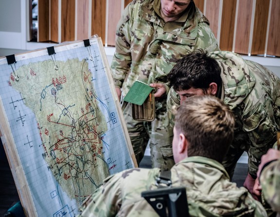 New Zealand Army uniformed soldiers look at a map. One has their back to the camera and has a weapon on the back.