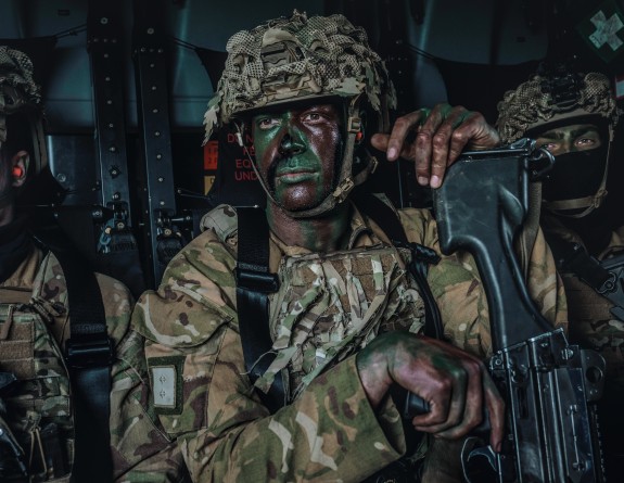 A New Zealand Army soldier in uniform, covered in camo paint looking to the left of the camera. They have a helmet on and are sitting strapped in with another soldier either side of them.